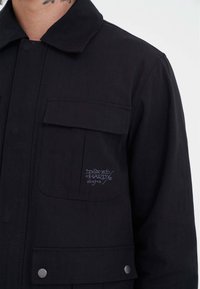 Czarna kurtka z kieszenią na piersi z zapięciem na guzik i haftowanym logo "Don Ed Hardy designs" z przodu po lewej stronie.