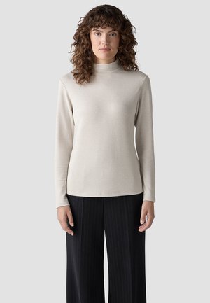Woman with curly hair wearing a beige long-sleeve mock neck top and black wide-leg pinstripe pants, standing against a plain background.