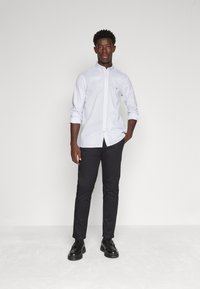 Camicia bianca a maniche lunghe con colletto abbottonato, indossata con pantaloni neri skinny e scarpe nere. Design semplice con vestibilità classica.