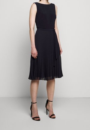 Femme portant une robe midi plissée noire sans manches et des sandales à talons hauts noires, debout contre un mur gris clair.