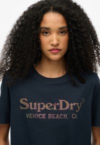 Marineblå bomulds t-shirt med et farverigt rhinsten-logo "SuperDry" og "VENICE BEACH, CA." på fronten, korte ærmer og rund halsudskæring.