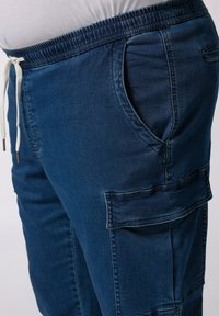 Dżinsowe spodnie dresowe w ciemnym niebieskim kolorze z elastycznym pasem, sznurkiem do ściągania, kieszeniami bocznymi oraz kieszenią cargo na nodze.