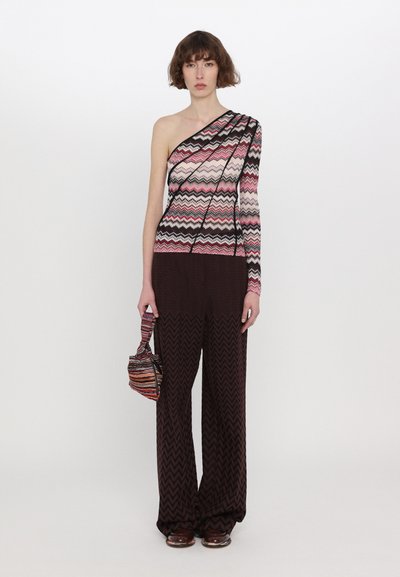 Asymmetrisches Oberteil mit einem Zickzackmuster in Pink, Schwarz und Weiß; kombiniert mit weiten braunen gemusterten Hosen; das Model hält eine kleine, mehrfarbige Tasche.