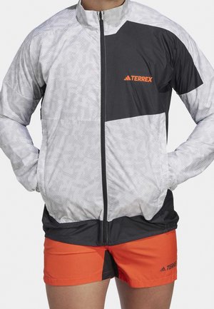 Personne portant une veste zippée Terrex grise et noire et un short Terrex orange vif avec les mains dans les poches de la veste.