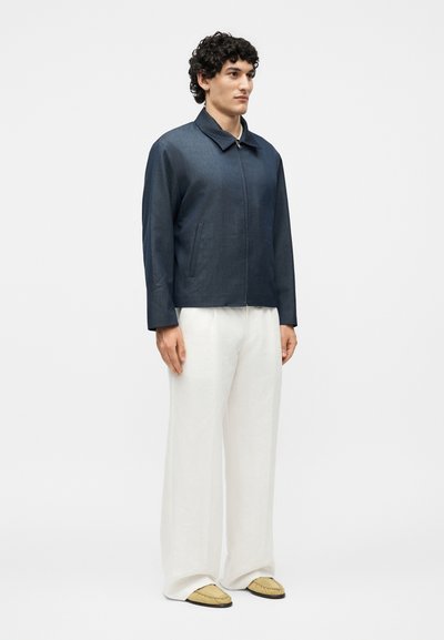 Άνδρας που φοράει σκούρο μπλε ζακέτα με φερμουάρ, φαρδιά λευκά παντελόνια και μπεζ παπούτσια χωρίς κορδόνια, μπροστά σε απλό φωτεινό φόντο.