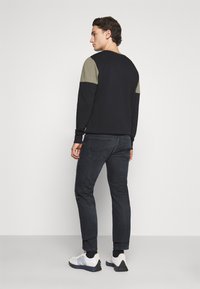 Moleton preto de gola redonda com painéis em verde-oliva nos ombros, combinado com jeans escuros e sapatilhas brancas, visto de costas.