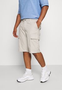Pantalones cortos cargo beige con bolsillos laterales, hechos de tela de algodón. Combinados con una camisa de color azul claro y zapatillas deportivas blancas, de pie sobre un fondo neutro.