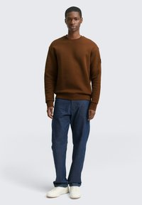 Felpa marrone con texture, colletto a giro e orlo a coste, abbinata a jeans blu scuro e sneakers bianche. Design minimalista, vestibilità casual.