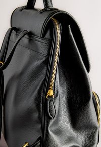 Schwarzer Leder-Rucksack mit strukturierter Oberfläche, der goldene Reißverschlussdetails und obere Griffe vor einem schlichten Hintergrund zeigt.