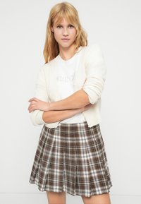 Κρεμ ζακέτα πάνω από λευκό T-shirt, συνδυασμένη με πλισέ καρό φούστα σε αποχρώσεις του καφέ και λευκού, με μοτίβο καρό πολλών χρωμάτων.