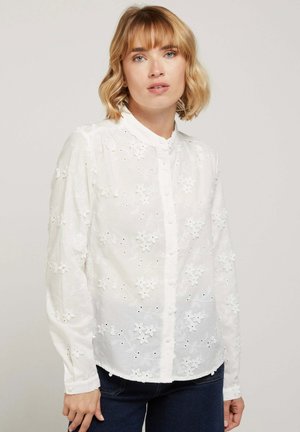 Chemise blanche à manches longues avec un col montant, ornée de broderies florales et de détails en œillets, fermeture à boutons sur le devant et texture légère.