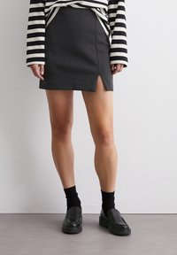 Zwarte minirok met een voorklep, gemaakt van gladde stof. Gepaard met een gestreepte langärmige top en zwarte loafers.