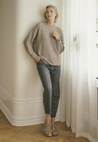 Beige textuur trui, grijze tapered broek en groene sandalen. De setting heeft zachte gordijnen en een houten vloer.