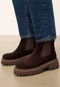 Rudos suede kulkšnies batai su elastingais šonais ir stora rudai guma padais, turinčiu tekstūruotą protektorių. Derinami su mėlynomis džinsinėmis kelnėmis.