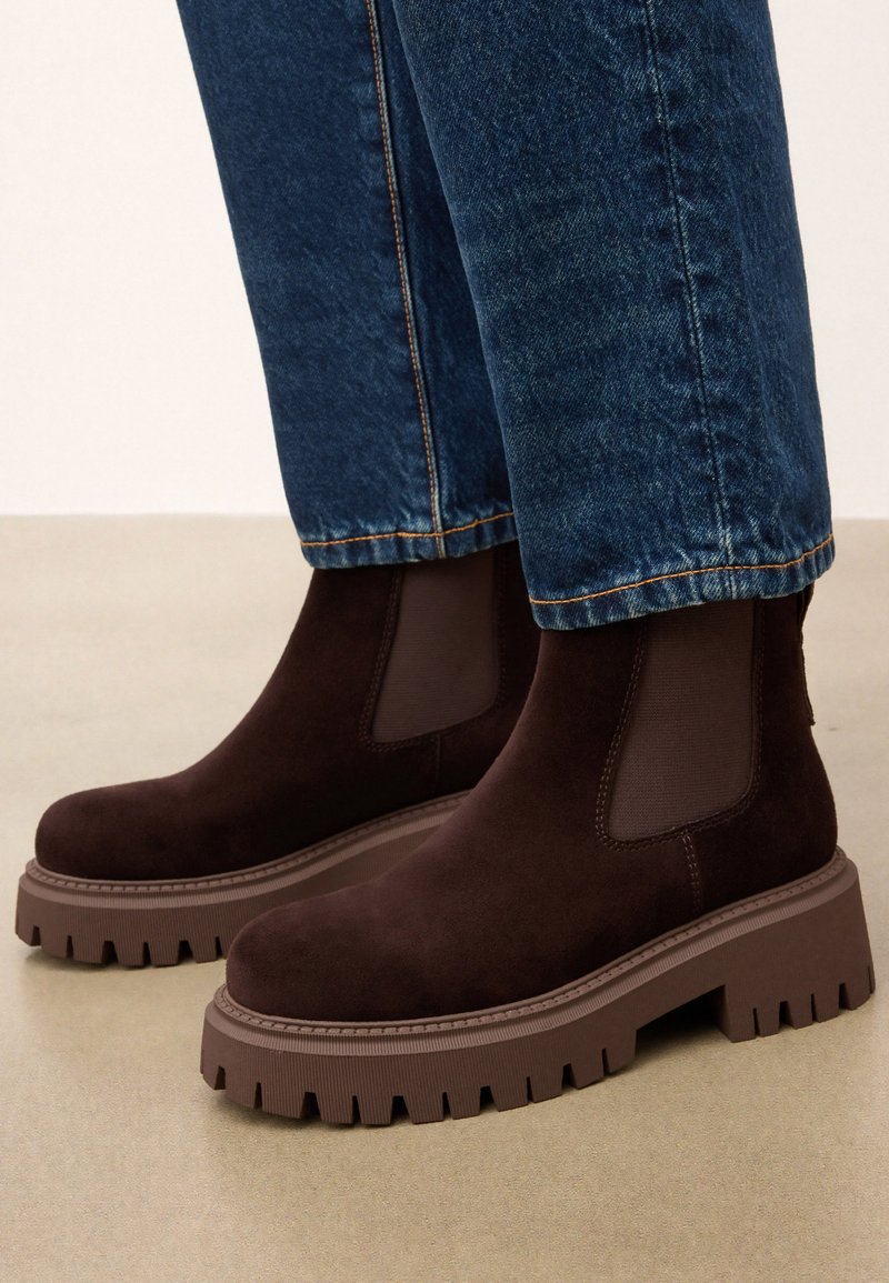 Rudos suede kulkšnies batai su elastingais šonais ir stora rudai guma padais, turinčiu tekstūruotą protektorių. Derinami su mėlynomis džinsinėmis kelnėmis.