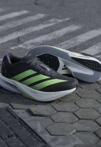 Μαύρα και πράσινα αθλητικά παπούτσια Adidas τοποθετημένα πάνω σε εξαγωνικά πλακάκια πεζοδρομίου με θολή διάβαση πεζών στο παρασκήνιο.