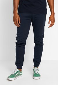 Marineblauwe tapsgewijze joggers van zachte stof met paneeldetails en ribgebreide boorden, gecombineerd met groen-witte sneakers met een low profile.