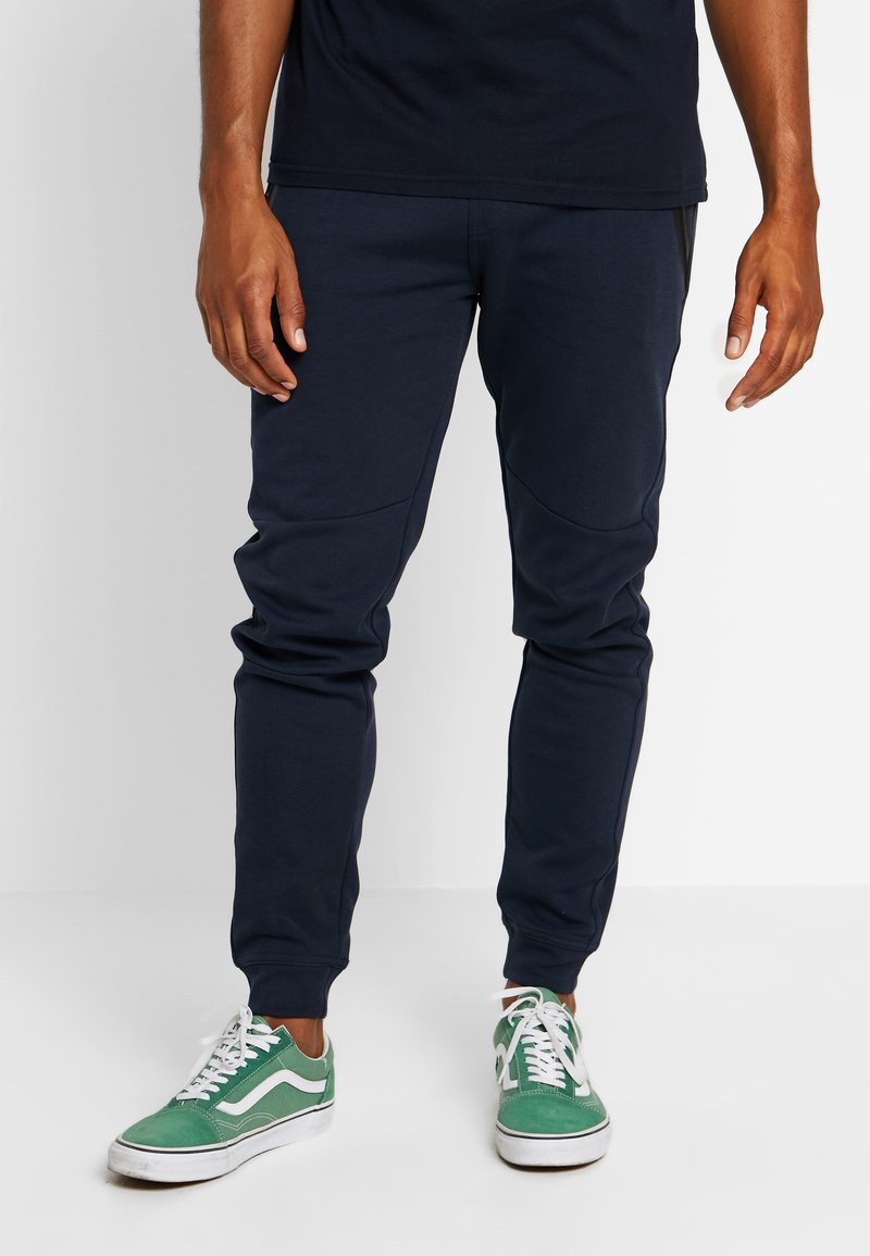 Marineblauwe tapsgewijze joggers van zachte stof met paneeldetails en ribgebreide boorden, gecombineerd met groen-witte sneakers met een low profile.