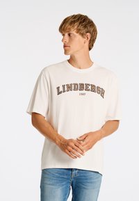 Camiseta de manga corta y cuello redondo en color blanco roto, con un estampado en relieve en negro y marrón que dice "LINDBERGH 1927" en el pecho.