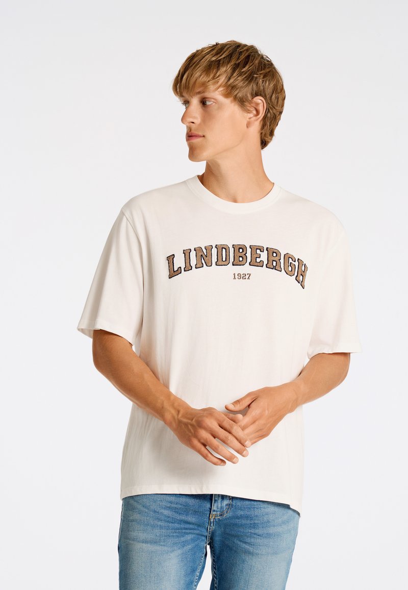 Camiseta de manga corta y cuello redondo en color blanco roto, con un estampado en relieve en negro y marrón que dice "LINDBERGH 1927" en el pecho.