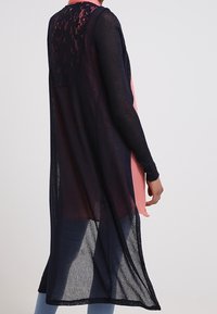 Lång, öppen cardigan i marinblått transparent material med en kontrasterande panel i rosa, spetsdetaljer på baksidan och långa ärmar.