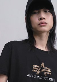 T-shirt noir à manches courtes avec un logo rehaussé de doré et d'argent. Le tissu semble lisse et la coupe est décontractée.