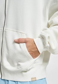 White zip-up hoodie with a relaxed fit, soft texture, front pockets, visible stitching, and a small label at the hem.