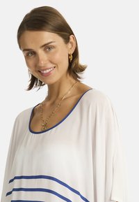 Giovane donna con capelli corti castani sorride, indossando un top bianco con bordi e strisce blu, orecchini a cerchio dorati e una collana a catena dorata.