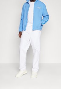 Cazadora cortavientos azul claro con un logo blanco, combinada con pantalones blancos sueltos y zapatillas blancas, mostrando un estilo deportivo casual.