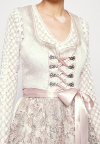 Robe de couleur claire avec des détails en dentelle complexe, broderies florales, quincaillerie en argent et un ruban en satin. Présente un décolleté en V profond et des manches longues.