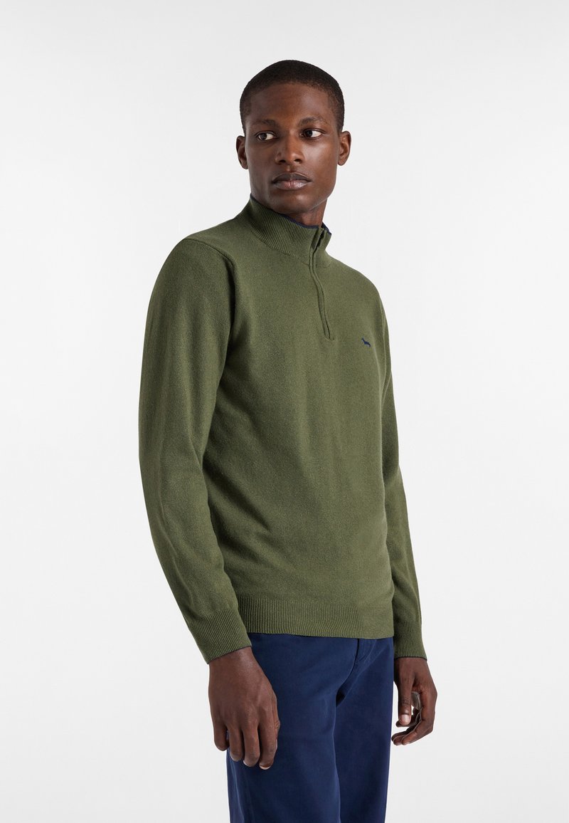 Zielony sweter z wysokim kołnierzem i pół zamkiem. Posiada ryflowane mankiety i dół. Subtelny detal z logo na klatce piersiowej. Noszony z granatowymi spodniami.