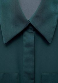 Chemise vert foncé avec un col large et pointu et une patte de boutonnage cachée, montrant la texture du tissu et les détails des coutures.