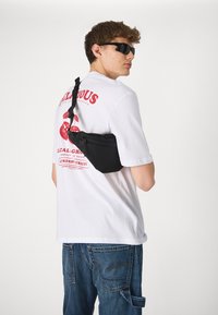 Bolso negro de hombro con cierre de cremallera, usado sobre una camisa blanca con diseño gráfico rojo. Unos jeans de mezclilla azul casual completan el look.