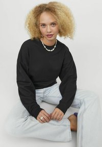 Felpa nera oversize con spalle scese, abbinata a jeans a gamba larga blu chiaro. Tra gli accessori degni di nota ci sono una collana di perle e un anello.