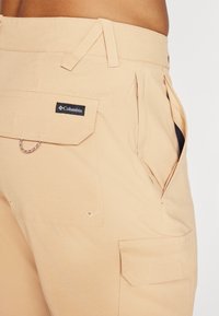 Beige cargobukser laget av lett stoff, med flere lommer, forsterkede sømmer og et logo-klistremerke på fronten.
