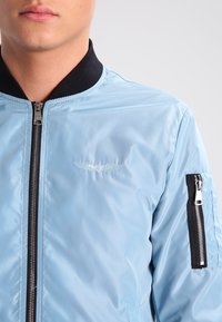 Světle modrá bomber bunda s lesklým povrchem, černý žebrovaný límec a kapsy na zip na rukávech. Obsahuje minimální branding.