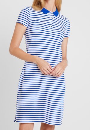Frau trägt ein weißes kurzärmeliges Kleid mit blauen horizontalen Streifen und blauem Kragen, Hände sanft vor dem Körper verschränkt.