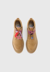 Stivaletti alla caviglia in suede tan con lacci colorati; i ciondoli decorativi includono fiori e una bicicletta. Dettagli delle cuciture attorno alla suola.