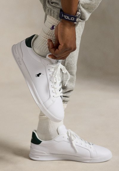 Inimene, kes kannab valgeid Polo tossusid ja valgeid sokke lillaka logoga, reguleerib ühe jalga kinga, riietatud heledatesse hallidesse dressipükstesse.