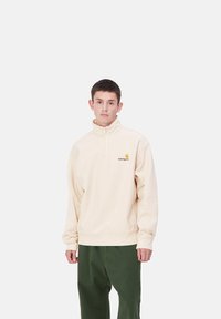Sudadera de poliéster beige con media cremallera y un logo en el pecho. Cuenta con un corte relajado, puños acanalados y cinturilla. Combinada con pantalones verdes.