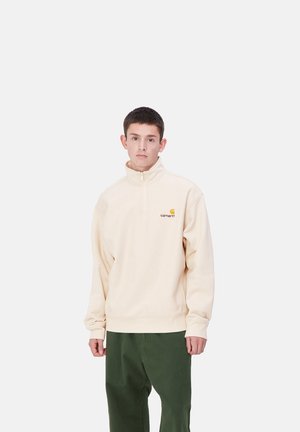 Sudadera de poliéster beige con media cremallera y un logo en el pecho. Cuenta con un corte relajado, puños acanalados y cinturilla. Combinada con pantalones verdes.