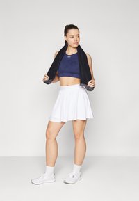 Sujeta de deporte azul marino, falda plisada blanca, zapatillas blancas y calcetines. Toalla negra caída sobre los hombros. Tela suave, estilo de ropa activa casual.