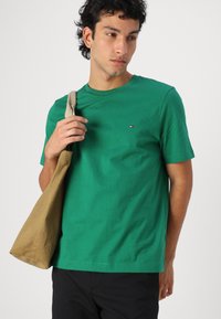 T-shirt en coton vert à col rond, avec des manches courtes et un petit logo sur la poitrine. Le modèle tient un sac fourre-tout marron.