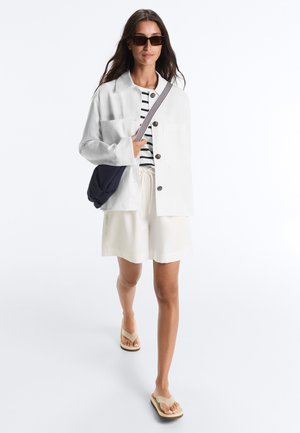 Femme portant des lunettes de soleil, une veste blanche, une chemise rayée, un short blanc, des sandales beiges et un sac bandoulière bleu marine, marchant vers l'avant.