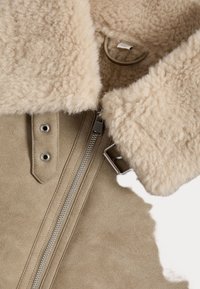 Beige schapenvachtjas met een pluizige kraag, zilveren rits en drukknopdetails op een zachte suede buitenkant. Gelaagd, warm en knus ontwerp.