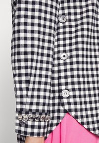 Chemise à carreaux vichy noir et blanc à manches longues, avec de gros boutons et un poignet orné de strass transparents. Tissu rose en dessous.