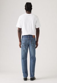 Jeans de mezclilla azul con corte recto, que cuentan con dos bolsillos traseros y un parche de cuero marrón en la cintura. Combinados con una camiseta blanca sin estampado.