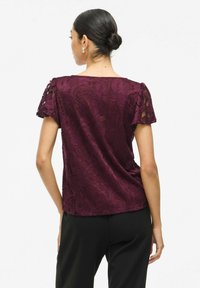 Top in pizzo borgogna con maniche corte e motivi floreali intricati; texture liscia e vestibilità comoda, indossato con pantaloni neri.