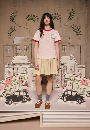 Junge Frau in pink gestreiftem Hemd, beigem Rock, grauen Socken und braunen Schuhen steht zwischen illustrierten Blumen, Autos und Häusern vor einem dekorativen Hintergrund.