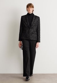 Zwarte gebloemde blazer met een notch-lapel, gecombineerd met een zwarte coltrui en een wijde broek. Model houdt een kleine metalen clutch vast.
